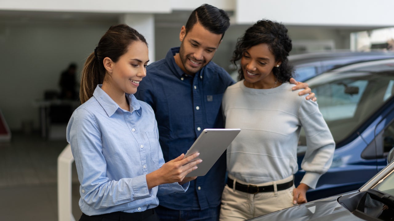 Saleswoman in blue shirt at a car dealership holds a tablet while talking to a smiling couple.