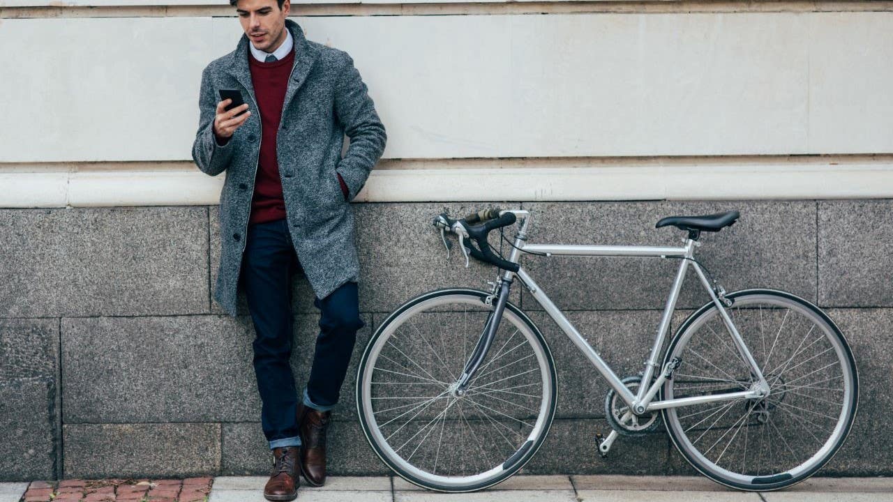 A man and a bicycle lean against the side of a building. The man is holding his phone and looking at it.