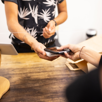 Close-up of a female customer in boutique making contactless mobile payment for her purchase. Customer holding cell phone over credit card reader at checkout counter with a sneeze guard in boutique.