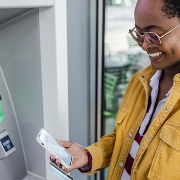 Young woman is holding a credit card and a mobile phone. She is standing in front of the ATM and thinking about the method of withdrawing money.
