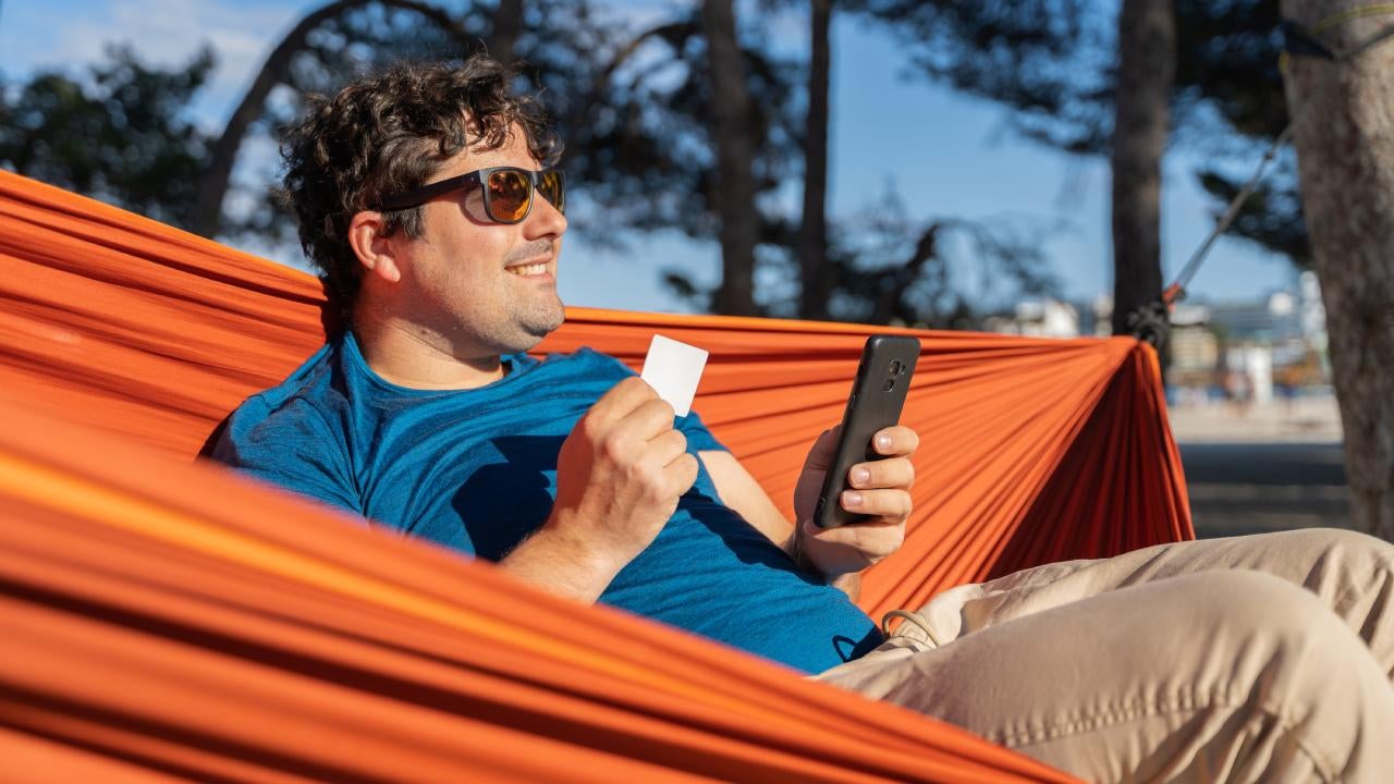 A portly white male wearing sunglasses is relaxing on his hammock outside, smiling in the sun holding a debit card in one hand and a smart phone in the other.
