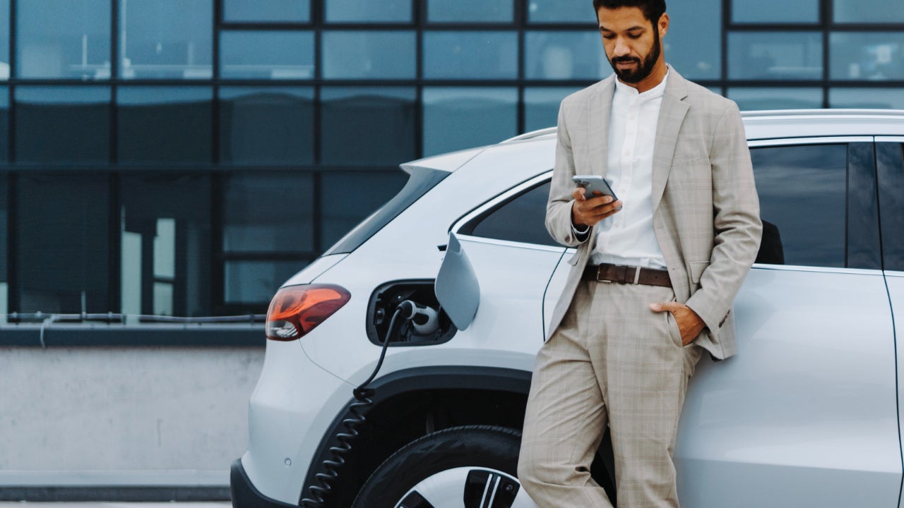 A young Black man in a suit uses his phone while charging an electric car.